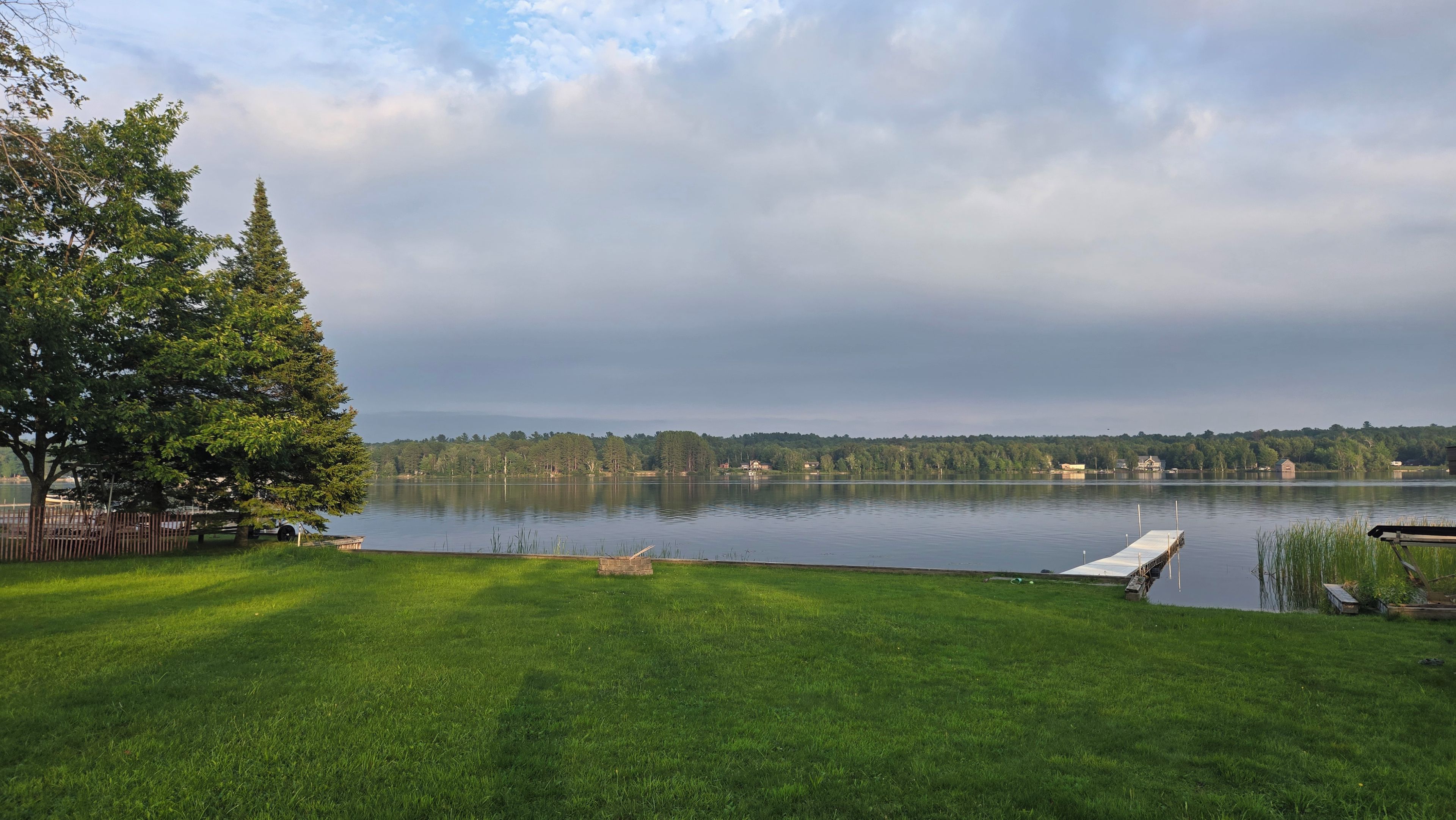 Une zone herbeuse avec une jetée qui s’avance sur un lac paisible, entourée d’arbres et bordée par une rive éloignée.