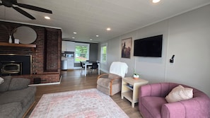 A living room with a fireplace, a dining area with a table and chairs, a television mounted on the wall, and a ceiling fan. - 3-bedroom • All season lakehouse (Hancock)