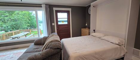 A bedroom with a bed, a sofa, a window with a view of greenery, and a door with a caution sign.