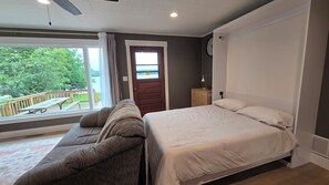 A bedroom with a bed, a sofa, a window with a view of greenery, and a door with a caution sign. - 3-bedroom • All season lakehouse (Hancock)