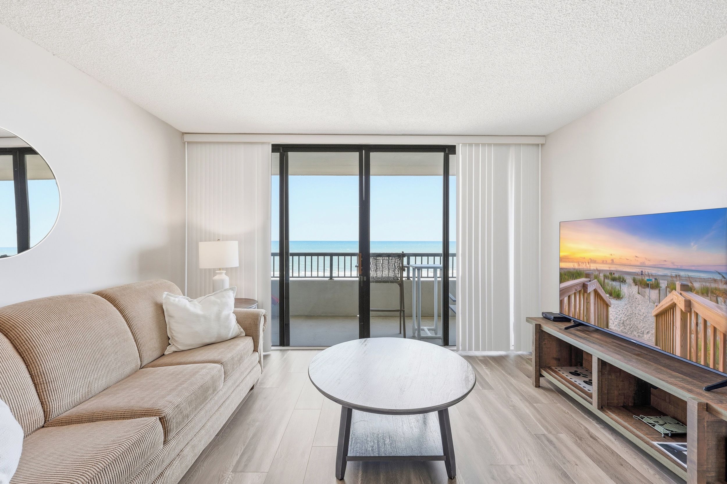 A hotel room with a sofa, a table, a TV, and a balcony with a view of the sea.
