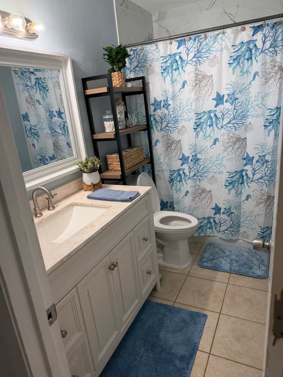 A bathroom with a sink, mirror, toilet, shower curtain, shelves, and a blue rug.