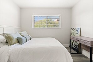 Une chambre à coucher avec un lit, une fenêtre avec des stores, une table de chevet et un coussin à motifs zébrés.