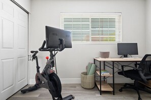 Un bureau à domicile équipé d’un vélo d’appartement, d’un bureau avec un ordinateur et d’une fenêtre avec des stores.