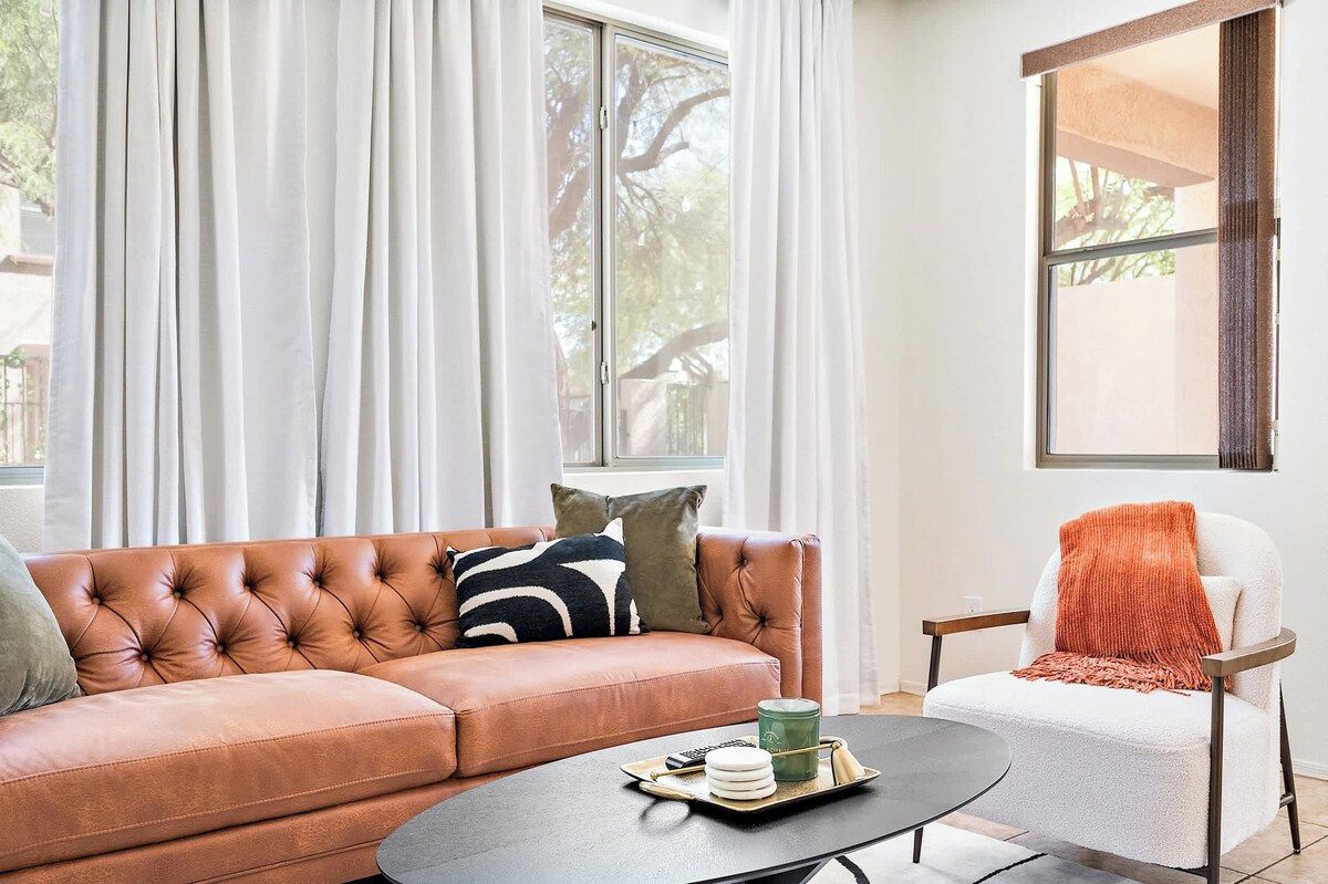 A living room with a brown leather sofa, a white armchair, a coffee table, and a window with curtains.