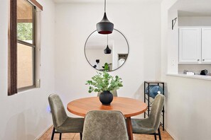 Ein Essbereich mit einem runden Holztisch, zwei gepolsterten Stühlen, einem Blumentopf, einer hängenden Pendelleuchte und einem wandmontierten Spiegel.