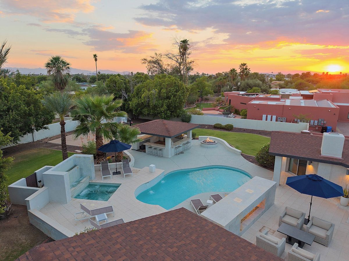 Una piscina con jacuzzi integrado, rodeada de sillas de descanso y sombrillas, con árboles y edificios al fondo durante el atardecer.