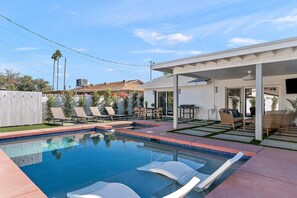 Ein Swimmingpool mit Liegestühlen, einer überdachten Terrasse und Blick auf benachbarte Häuser.