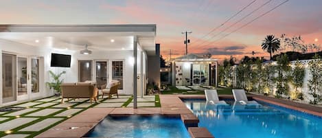 A modern outdoor pool area with a clear sky, a covered seating area, and a well-maintained lawn.