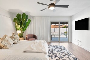 Un dormitorio con una cama, ventilador de techo, una planta grande, una silla y vista a través de un ventanal.