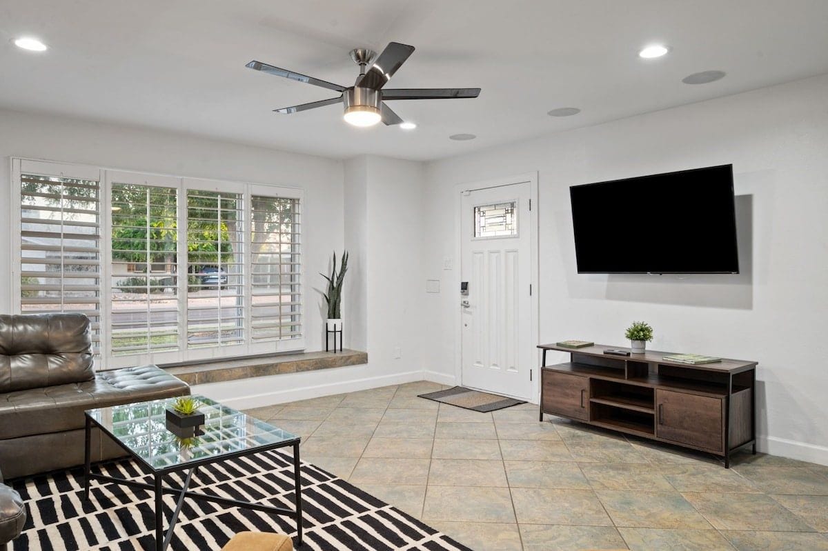 A modern living room with a leather sofa, a glass coffee table, a TV mounted on the wall, and a ceiling fan.