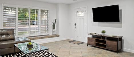 A modern living room with a leather sofa, a glass coffee table, a TV mounted on the wall, and a ceiling fan.