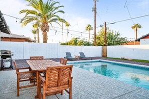 Un espace aménagé au bord de la piscine, avec une table et des chaises en bois, des chaises longues et un barbecue.