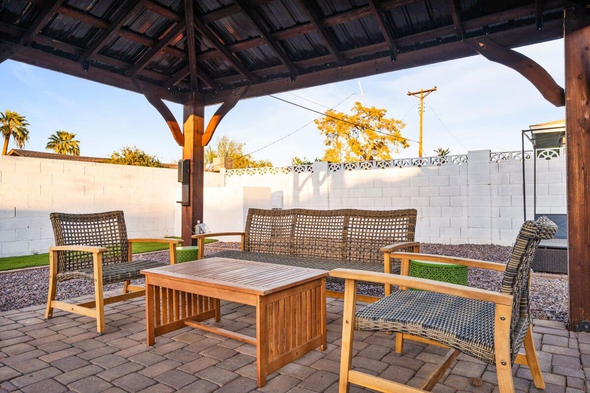 En udendørs terrasse med et træbord og fletstole, en pergola over hovedet og en hvid væg med et dekorativt hegn.