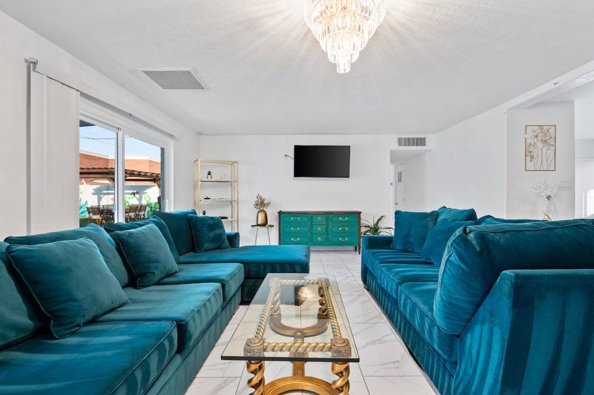 A modern living room with teal sofas, a glass coffee table, a chandelier, and a flat-screen TV on the wall.