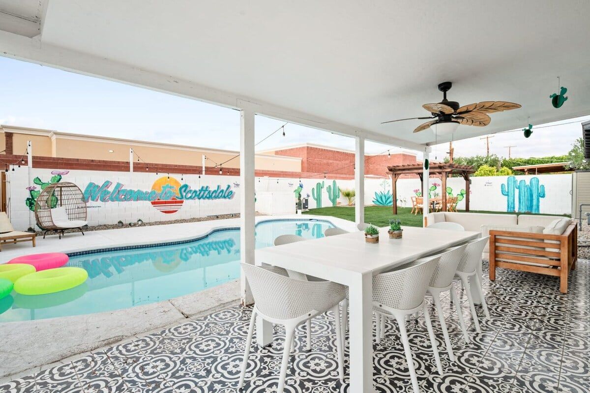 Un espace aménagé au bord de la piscine, avec une table et des chaises blanches, un ventilateur de plafond et une fresque murale.