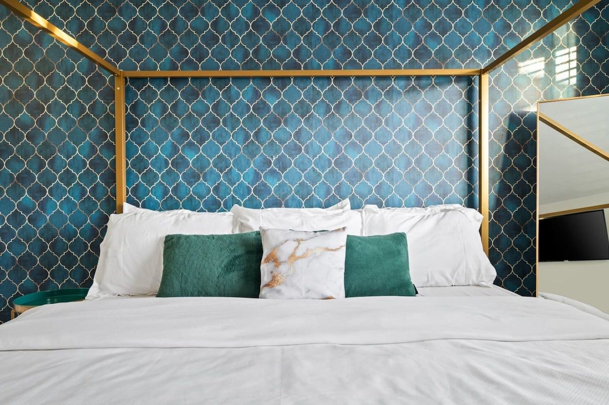 A bed with white bedding and green pillows, a gold-framed headboard, and a wall with a geometric pattern.