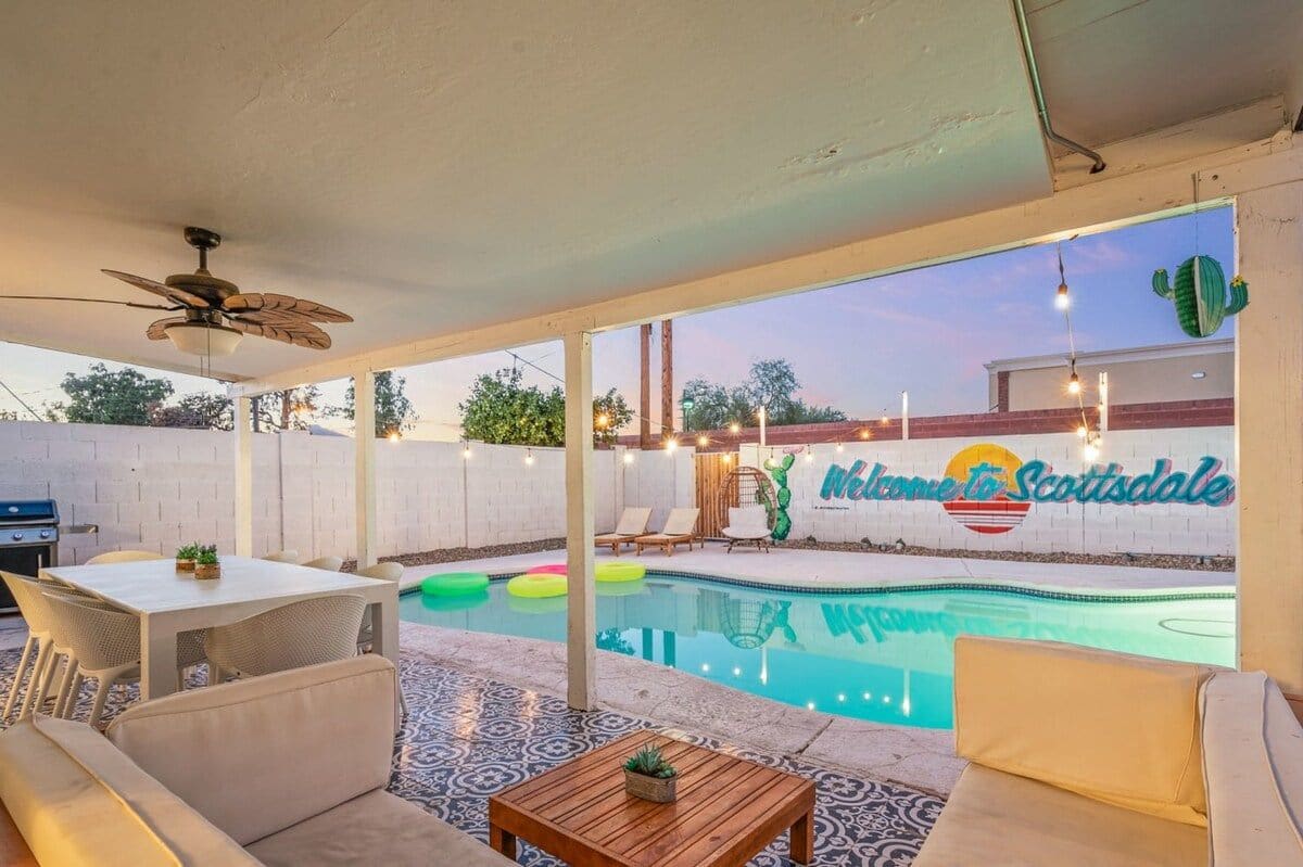 Un área de piscina exterior con ventilador de techo, mesa y asientos de descanso.