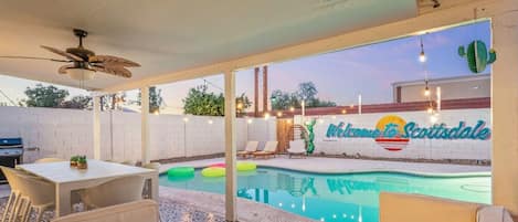 Un área de piscina exterior con ventilador de techo, mesa y asientos de descanso.