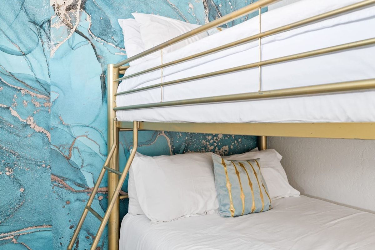 Uma cama beliche com roupa de cama branca e estrutura dourada, em frente a uma parede verde-água texturizada.