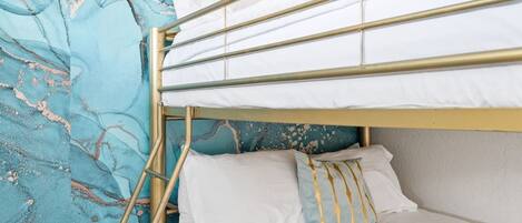 Una cama litera con ropa blanca y estructura dorada, frente a una pared azul verdosa con textura.