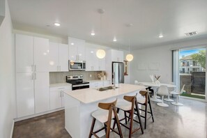 A modern kitchen with white cabinetry, a central island with bar stools, and a dining area with white chairs. - Chandler Townhome 1 🌵 Walk to Downtown Chandler, Community Pool & Grill (Chandler)
