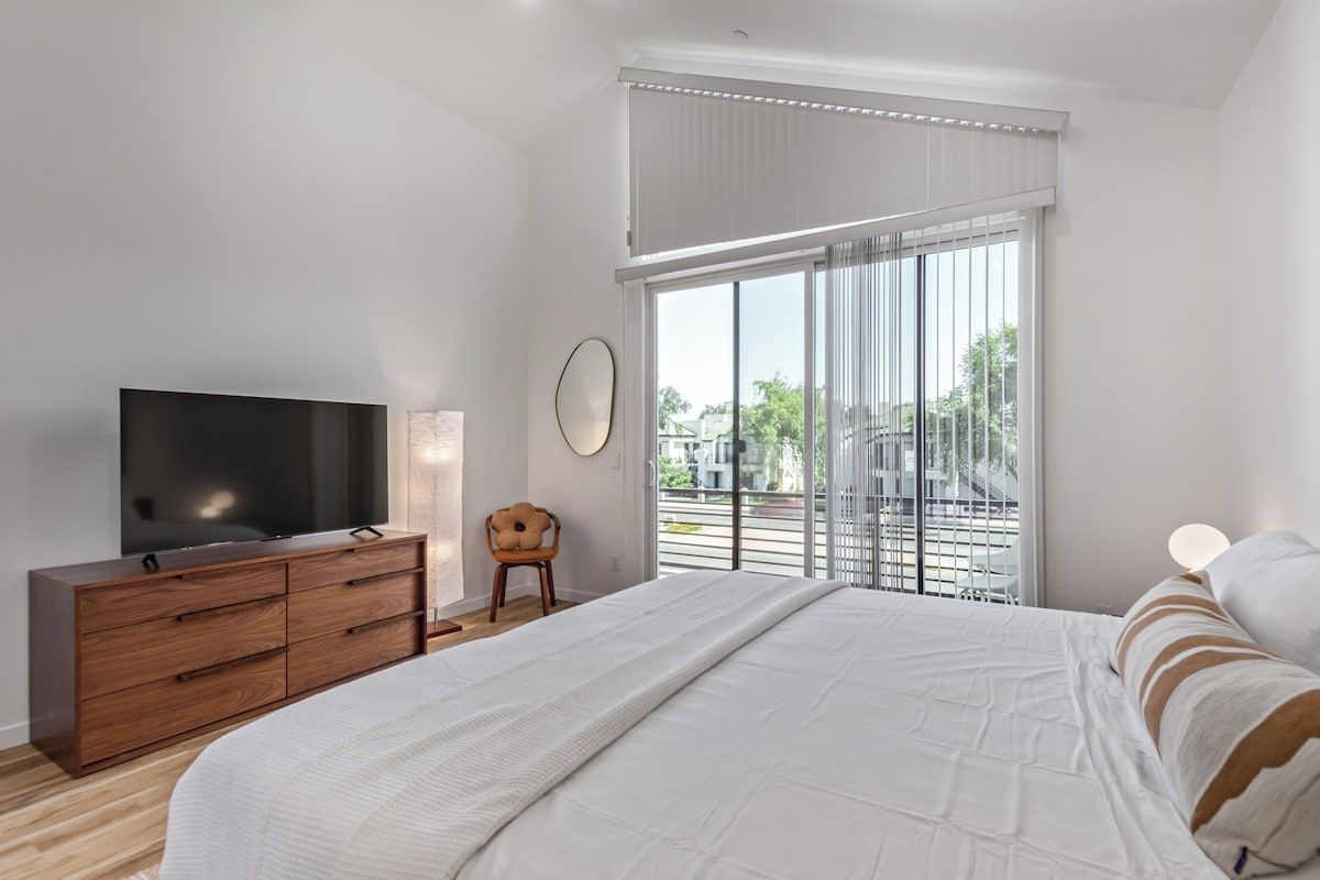 Un dormitorio con una cama, un televisor sobre un aparador de madera, una silla y un ventanal con persianas.