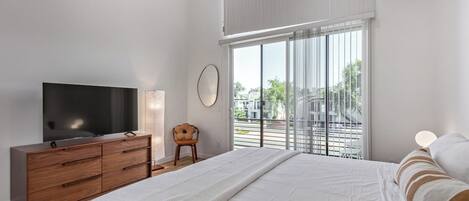 A bedroom with a bed, a TV on a wooden dresser, a chair, and a window with blinds.