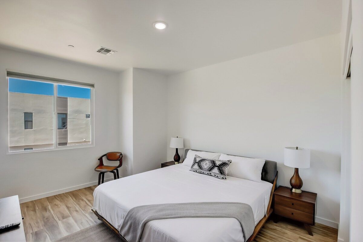 Une chambre à coucher comprenant un lit, deux tables de chevet avec des lampes, une chaise, une fenêtre avec des stores et un bureau avec une chaise.