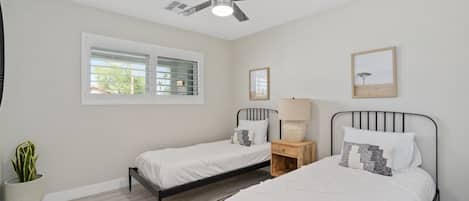 A bedroom with two single beds, a ceiling fan, a window with blinds, a bedside table with a lamp, and a framed picture on the wall.