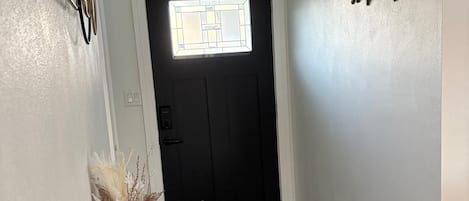 Um corredor com piso de madeira escura, uma porta branca com janela e uma pequena mesa com um vaso e um livro.
