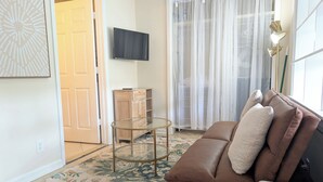 Een woonkamer met een bruine leren bank, een glazen salontafel, een flatscreen televisie, een houten kast en een groot raam met dunne gordijnen.