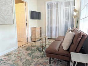 Ein Wohnzimmer mit braunem Ledersofa, einem Glastisch, einem Flachbildfernseher, einer Holzkommode und einem großen Fenster mit durchsichtigen Vorhängen.