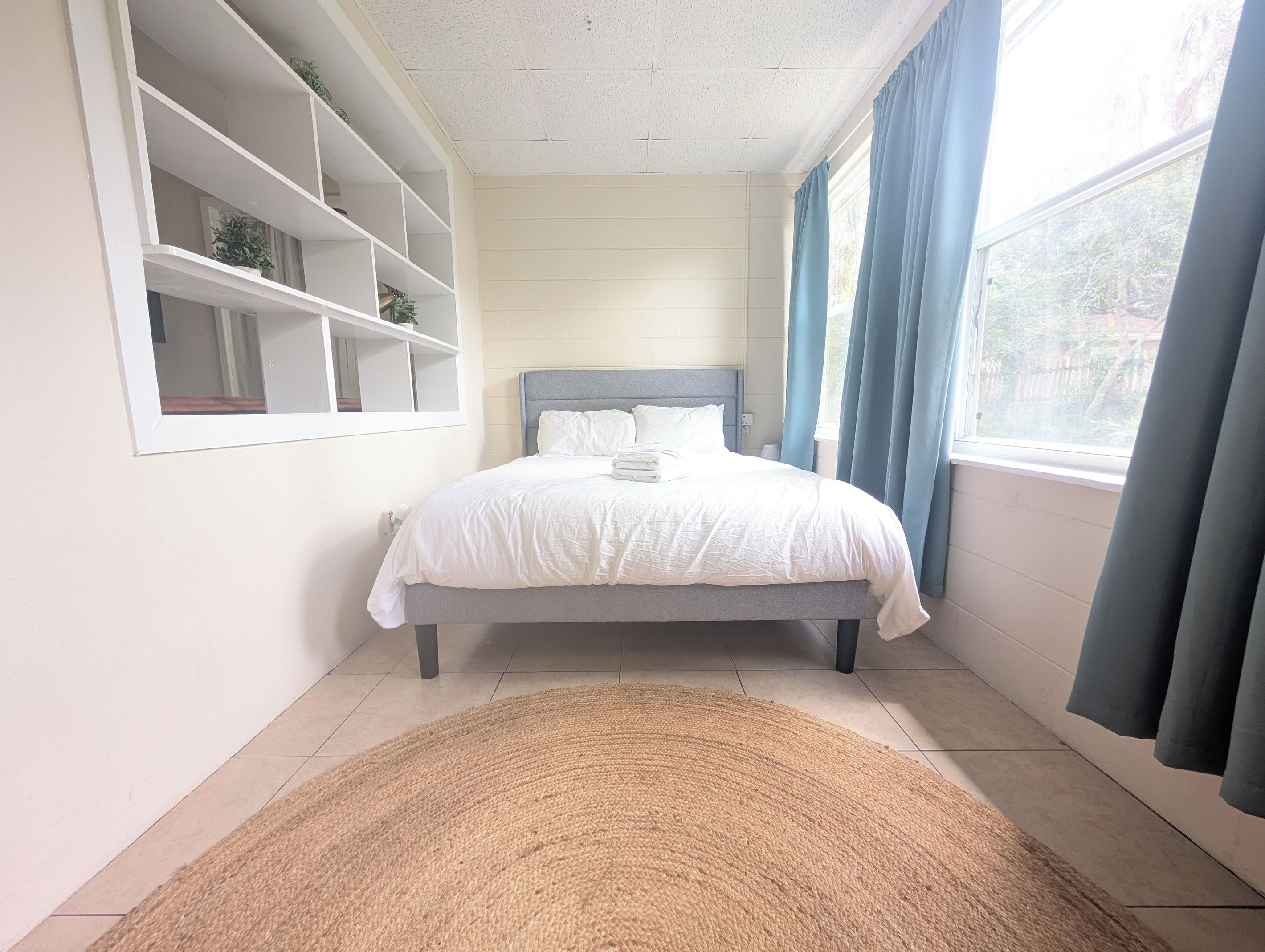 Un dormitorio con una cama, una ventana con cortinas, una estantería y una alfombra.