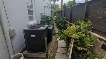 Een woonwijk met een smal pad, buitenunits voor airconditioning, een witte emmer en verschillende planten.