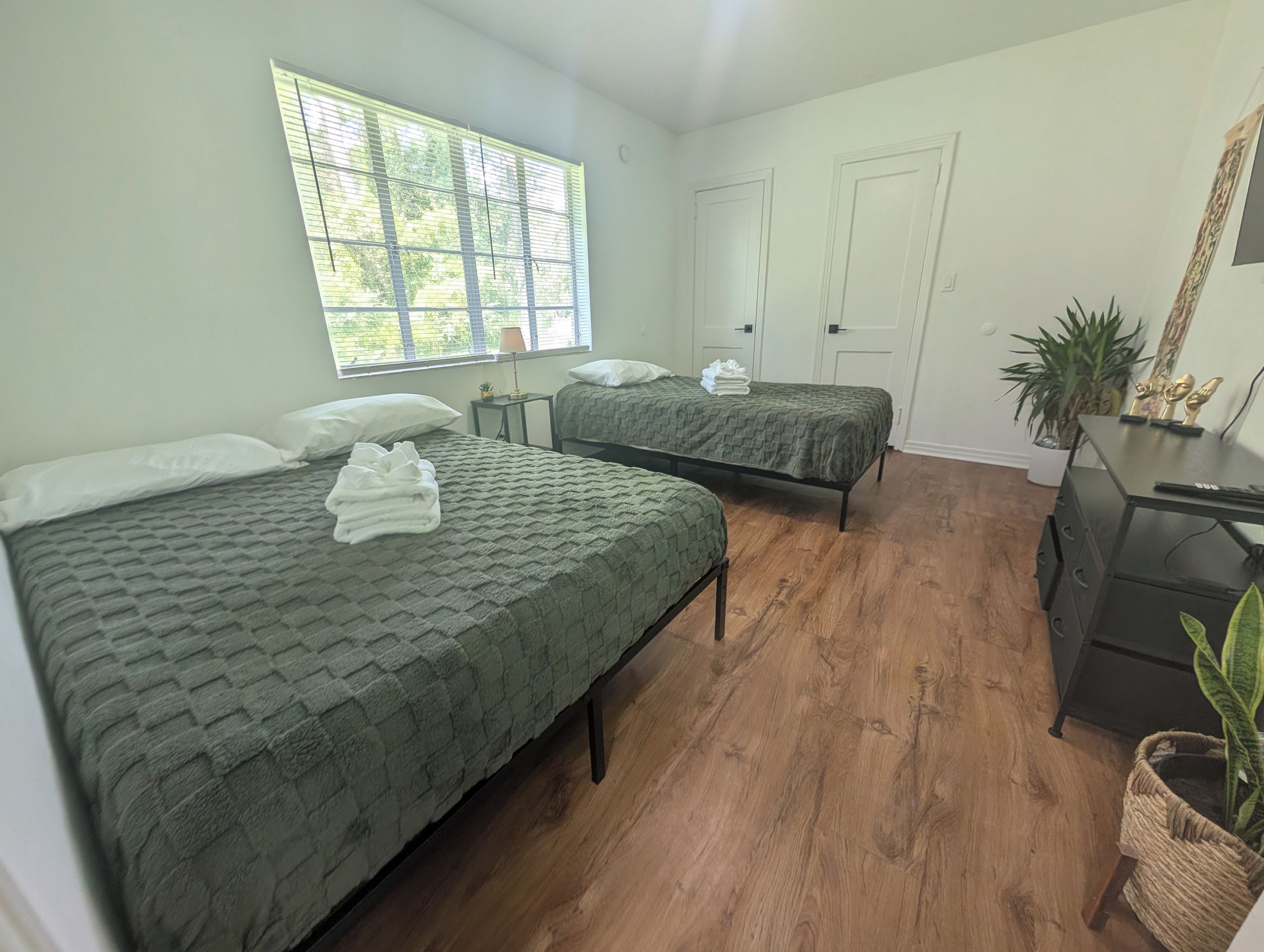 A room with two beds, a desk, a chair, a lamp, and a window with blinds.
