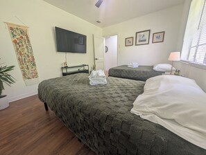 Habitación de hotel con dos camas, un televisor de pantalla plana, un tapiz en la pared y una ventana con persianas.