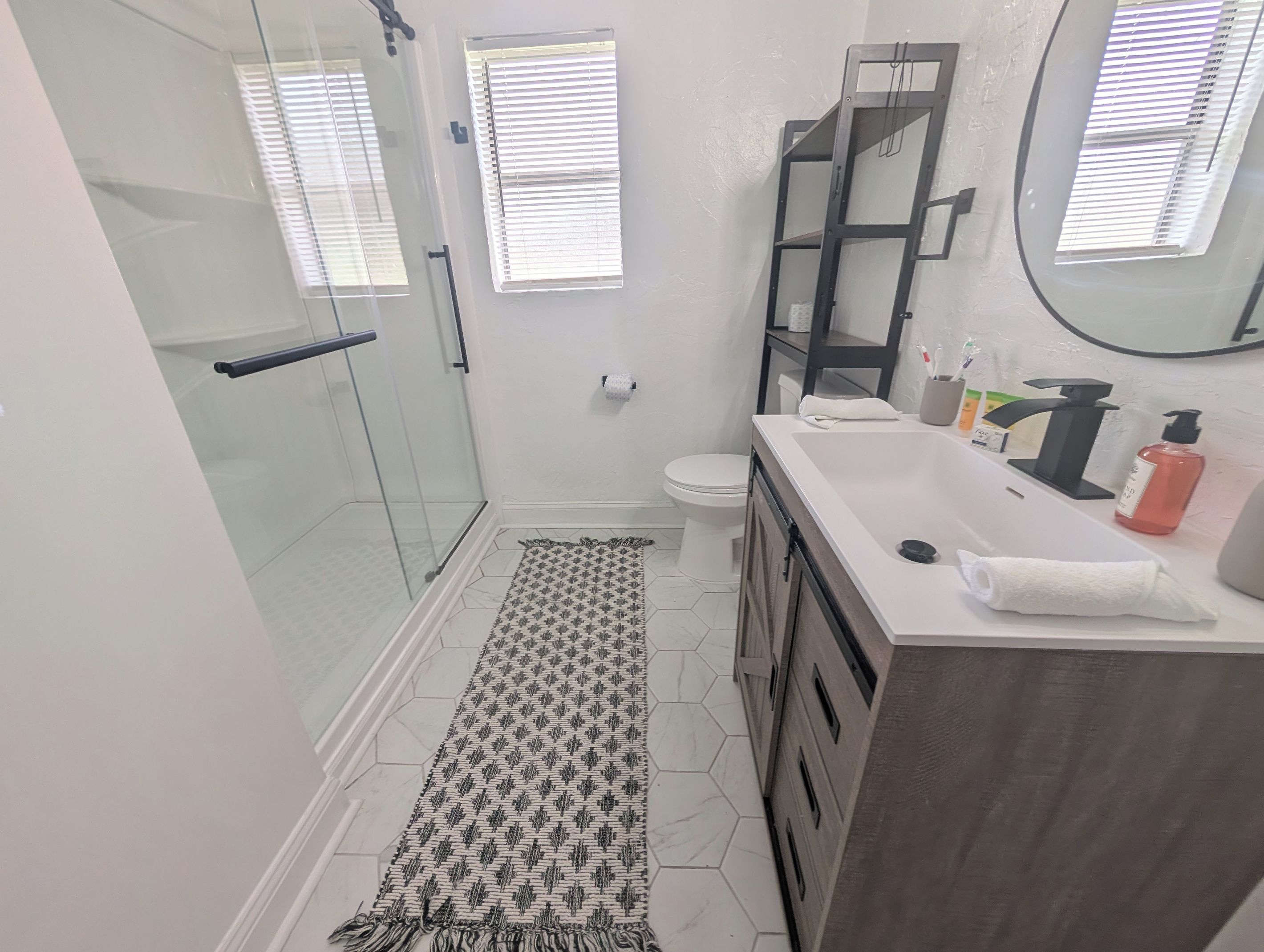 A modern bathroom with a glass shower enclosure, a white toilet, a vanity with a mirror, and a patterned rug on the floor.