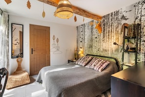 Quarto com cama, porta de madeira, parede com desenhos de folhas, luminária pendente e estante com plantas.