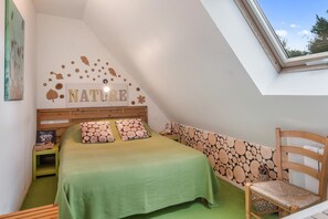 Quarto com teto inclinado, cama com roupa de cama verde, cabeceira de madeira, parede com decoração inspirada na natureza e uma janela.