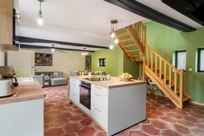 Eine Küche mit einer zentralen Kochinsel, ein Essbereich mit einer Couch und Stühlen sowie eine Holztreppe.