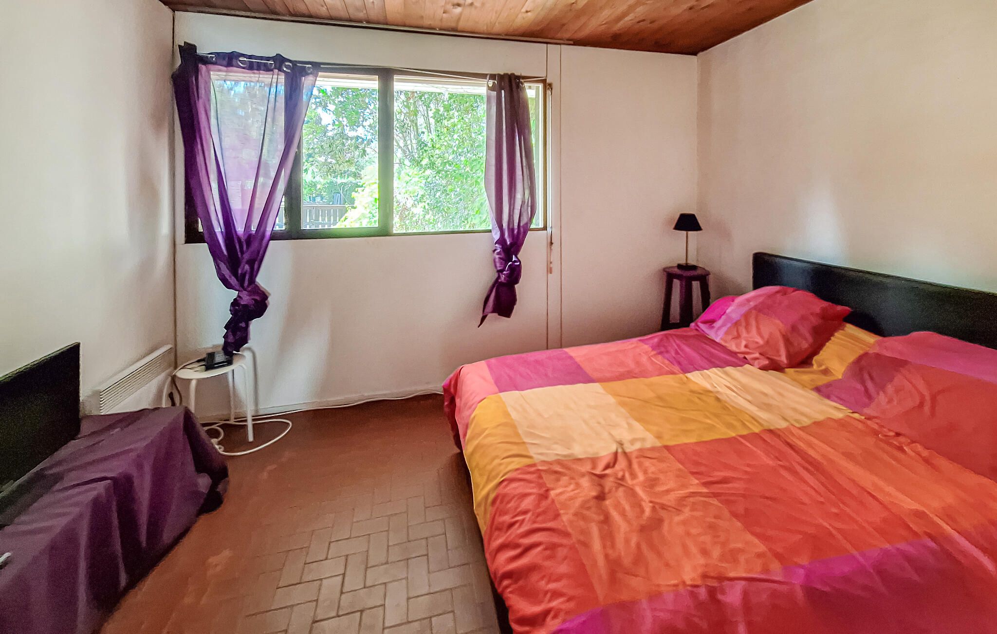 A bedroom with a bed, a window with curtains, a bedside lamp, and a small stool.