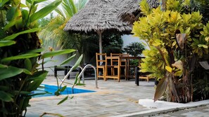 A pool area with a thatched roof structure, wooden furniture, and lush greenery.