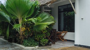 A patio area with a wooden table and chairs, surrounded by lush greenery including palm trees and various plants.