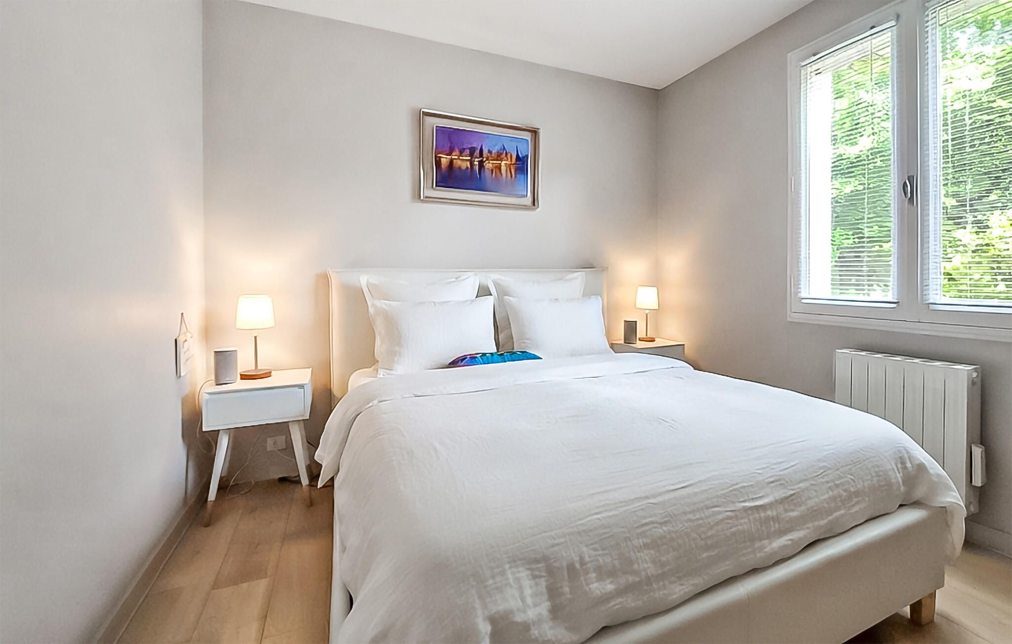 A bedroom with a large bed, two bedside tables with lamps, a framed picture on the wall, and a window with blinds.