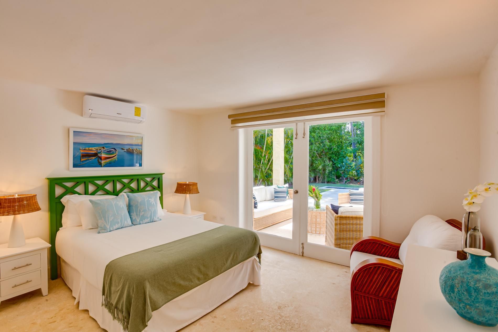 A bedroom with a bed, nightstands, a green headboard, a window with a view of greenery, and a white wall with a painting.