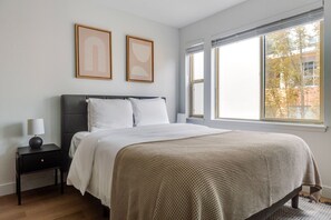 Ein Schlafzimmer mit einem Bett, einem Nachttisch, einer Lampe, zwei gerahmten Bildern und einem Fenster mit Jalousien.