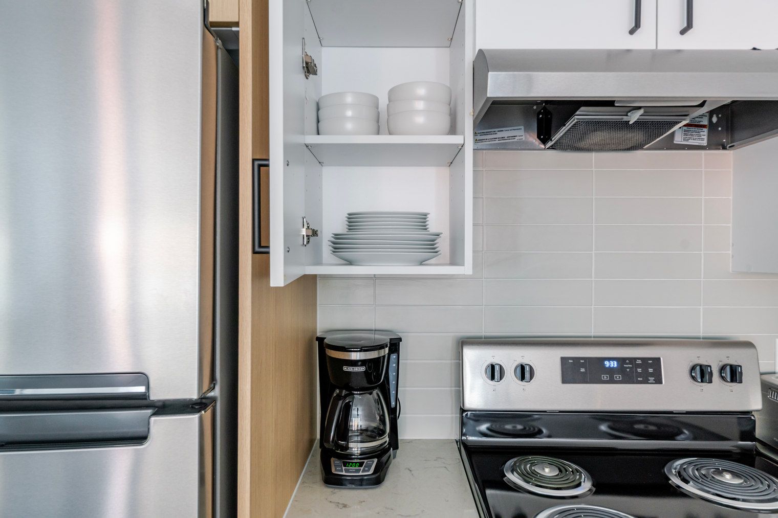 A compact kitchen with a stainless steel refrigerator, a coffee maker, a stove, and white cabinets with dishes.