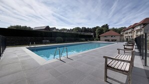 En rektangulær swimmingpool med en metalstige, omgivet af en fliseterrasse. Der er træbænke langs poolkanten.