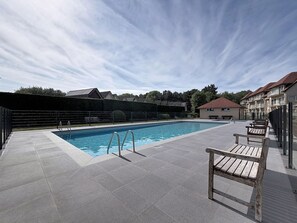 Una piscina rectangular amb una escala metàl·lica, envoltada per un solàrium emblamat. Hi ha bancs de fusta al costat de la piscina.